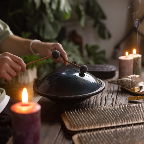 Workshop e Masterclass 14 Primo piano delle mani di una persona mentre suona un handpan con due battenti, circondata da candele e strumenti sonori in un’atmosfera rilassante dedicata al bagno sonoro armonico vibrazionale.