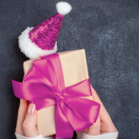 primo piano su due mani di donna che tengono un pacchetto regalo accandto al quele si trova un piccolo cappello da babbo natale rivestito di paillettes